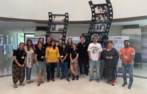A group of students and Dr. Roma Sur stand in front of a sculpture resembling a reel of film. 