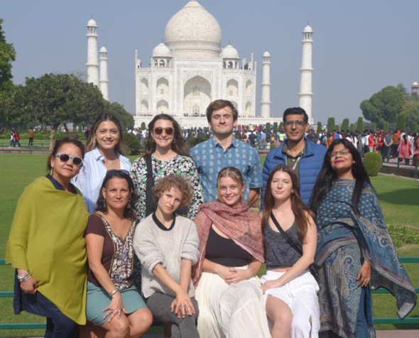 A group of students pose as a group in casual attire. They stand in front of the Taj Mahal, which is fully visible. 