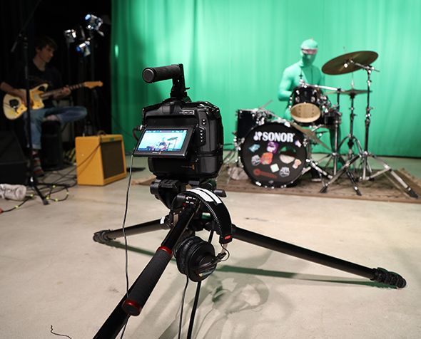 A guitarist and a drummer in a green jumpsuit play in front of the MFJS greenscreen. A camera on a tripod faces them. 