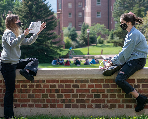 Two students wearing masks sit on campus to discuss a project