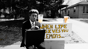 Black and white still image from Lemonade. A young black man in a suit and holding a briefcase sits in front of a lemonade stand. The lemonade in its pitcher and the letters on the sign on the stand are yellow, and the sign reads "When life gives you lemons. . ." 