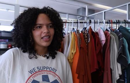 A young Black woman with curly hair poised to speak stands next to a rack of thrifted clothing 