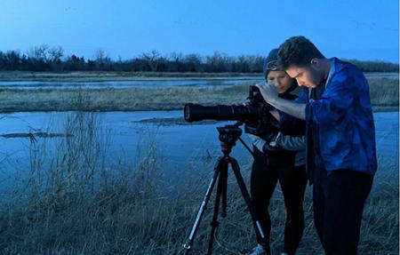 Two students adjust a camera on a tripod by a lake just before sunrise.