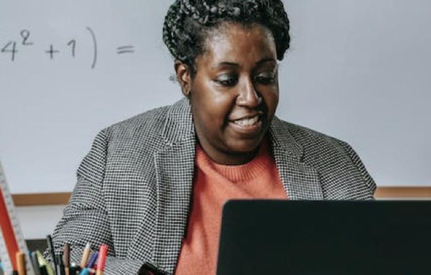 A black woman works on a laptop in front of a whiteboard with equations on it.