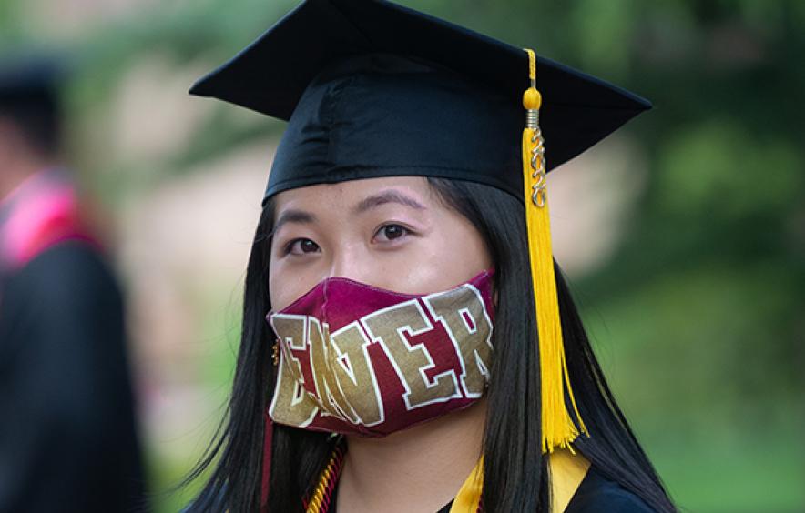 student wearing cap, gown, and DU face mask at commencement ceremony
