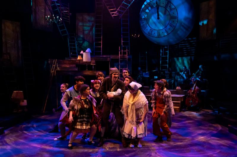 The cast of "Into the Woods" huddles in the center of a colorfully-lit stage. 
