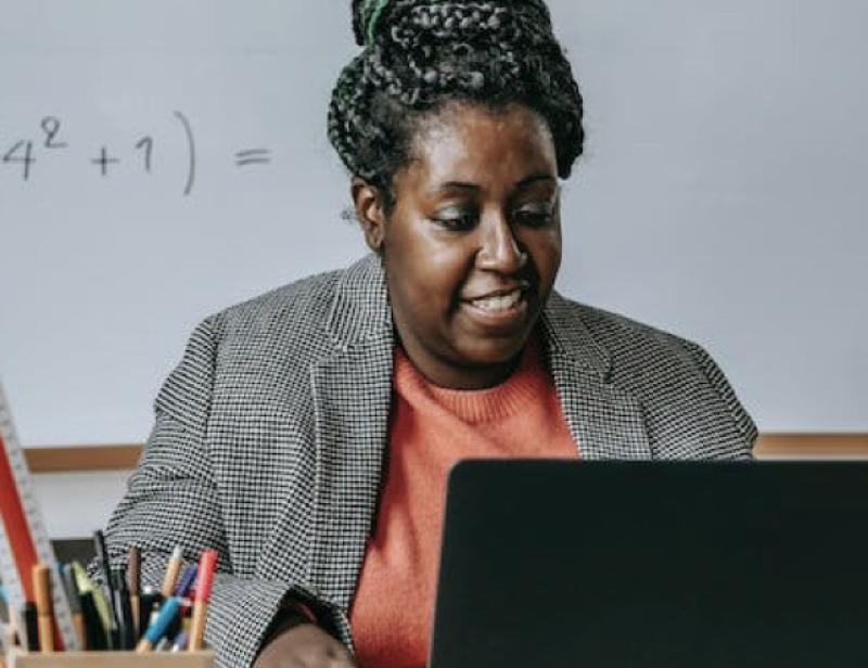 A black woman works on a laptop in front of a whiteboard with equations on it.