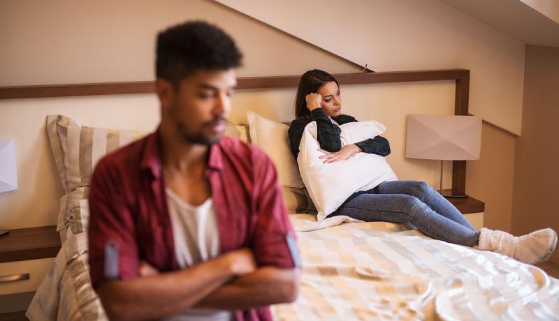 A couple sits on a bed turned away from each other, disgruntled.