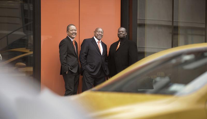 The Kenny Barron Trio stands against a wall with rushing traffic blurred in the foreground.