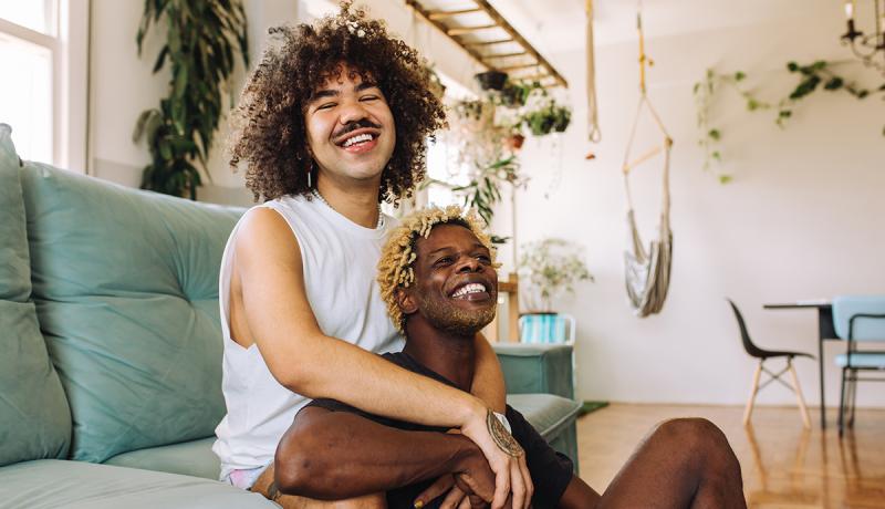 One man sits on a couch, embracing a second man who is sitting in front of him on the floor. The two are smiling comfortably and affectionately.