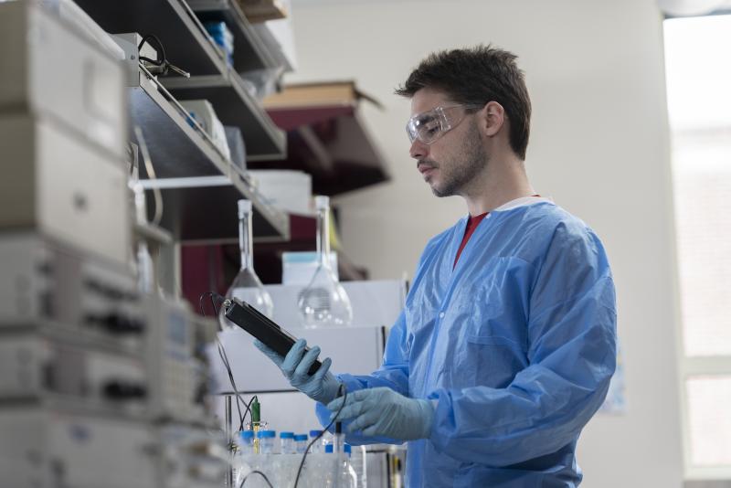 Image of a young man in blue lab scrubs looking at a small handheld machine in a lab.