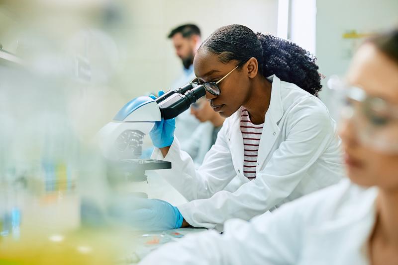 A Black woman in a lab coat peers through the lens of a microscope in a lab.