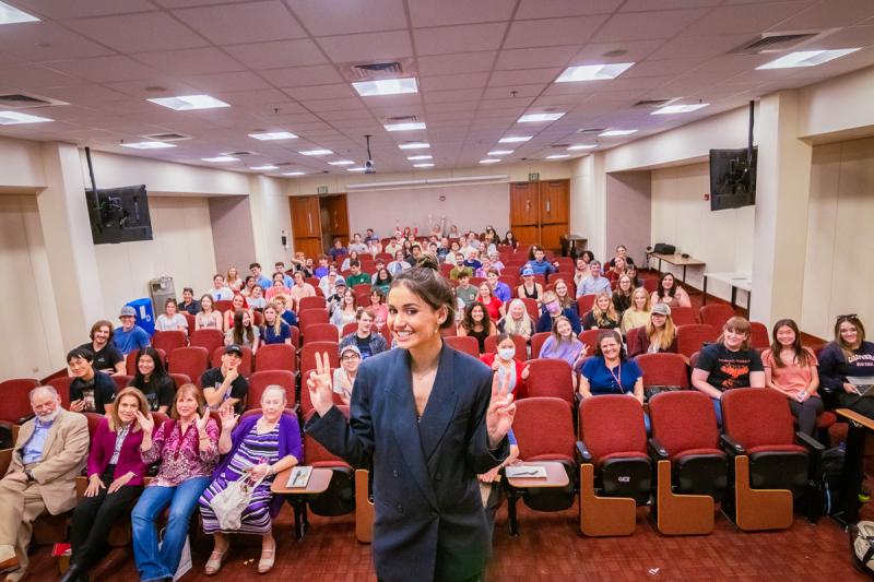 Sophia Kianni stands in front of a filled lecture hall as she and the audience hold up peace signs