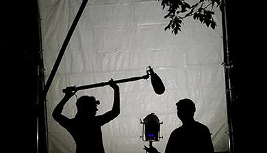 Two students stand silhouetted in the dark behind a backlit screen. One student holds a boom mic over the head of another student, who is standing next to a video camera.