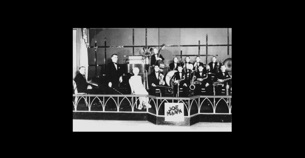 Black and white photograph of Joe Mann orchestra in the 1920s; all men are in black suits and there is a woman in white sitting in the front of the orchestra. 