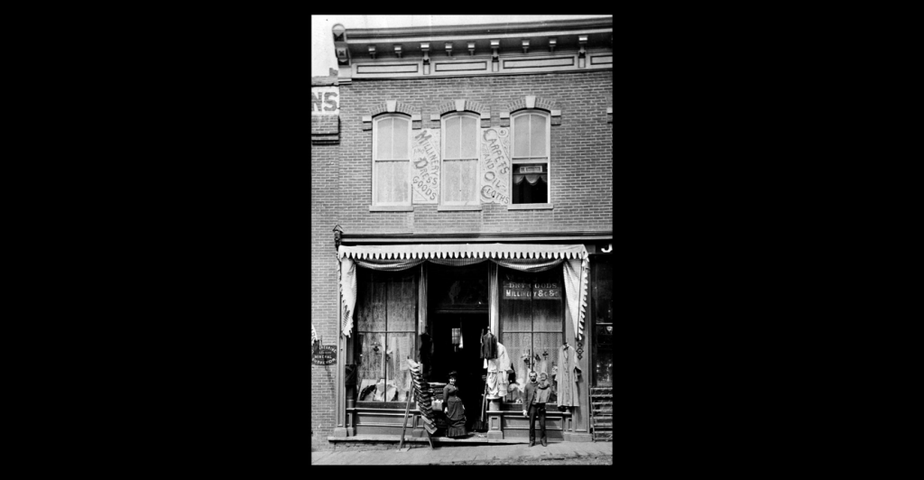 Black and white photograph of Rachofsky's store in Black Hawk late 1800s; there is a woman in a long dress with bustle and a man holding a baby standing in front of the store