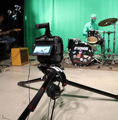 A guitarist and a drummer in a green jumpsuit play in front of the MFJS greenscreen. A camera on a tripod faces them.