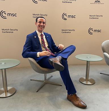 DU alumnus, Derek Dash, sits in front of a wall that reads "Munich Security Conference"