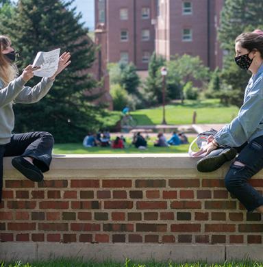 Two students sit outside on campus discussing a project while wearing face masks