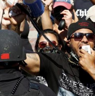 A group of people stand in protest against a line of police in riot gear. One man near the front is speaking into a bullhorn.