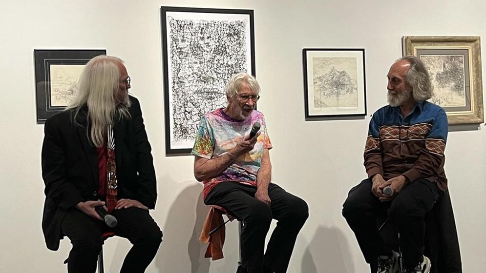 three older men seated on stools in front of paintings with one speaking into a microphone