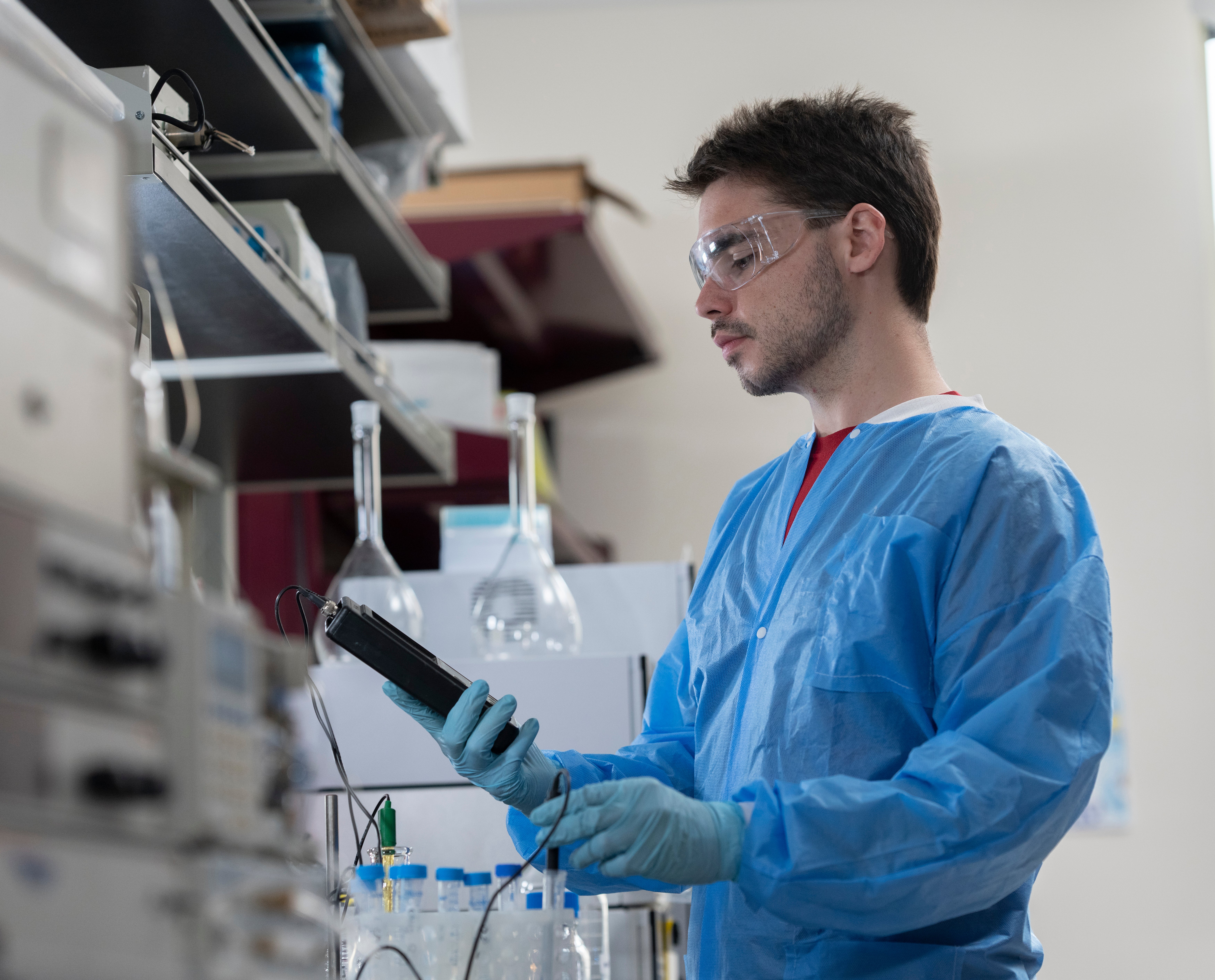 Image of a young man in blue lab scrubs looking at a small handheld machine in a lab.