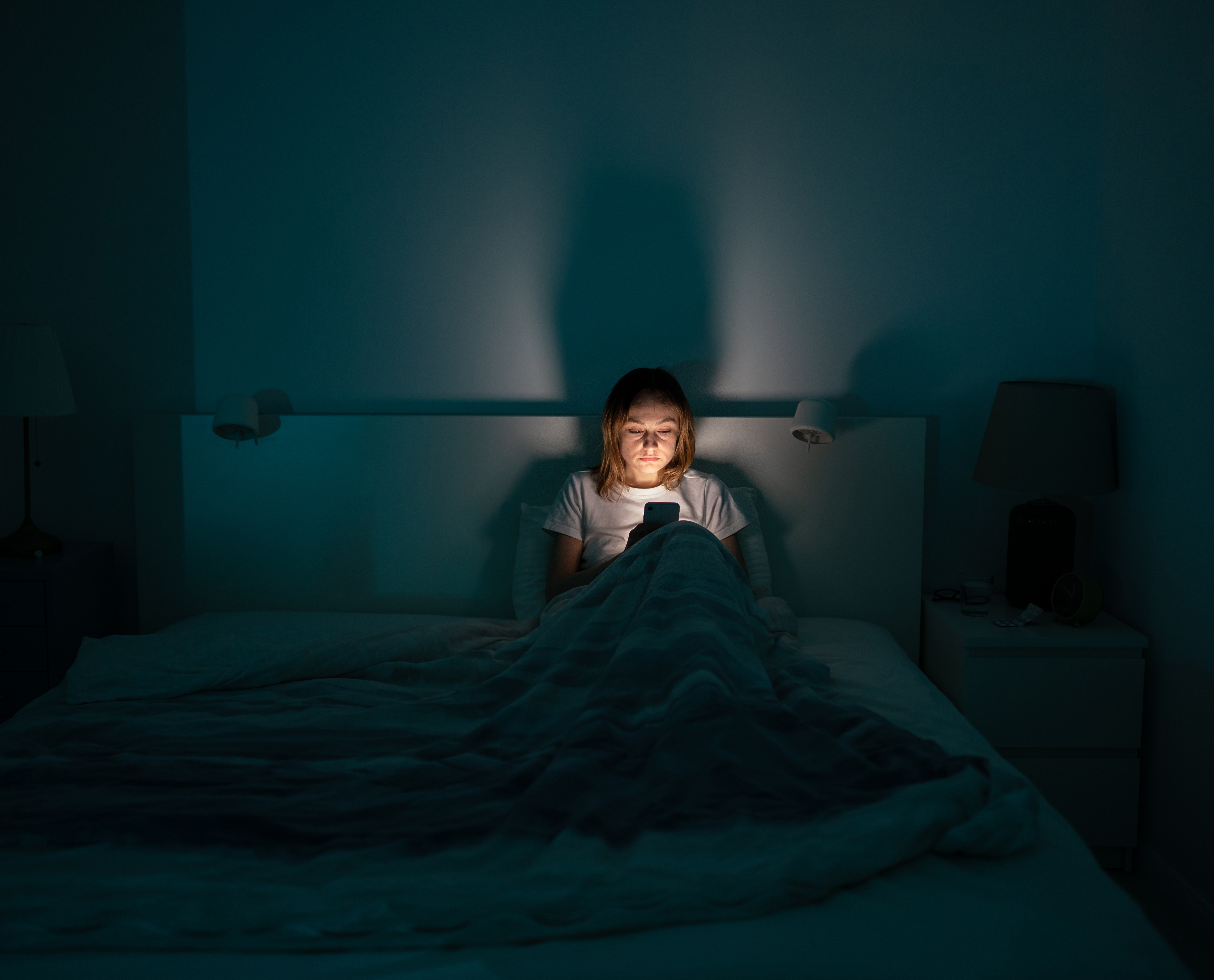A woman sits alone in a large bed on her phone with the lights off, the light from her phone illuminating her face.