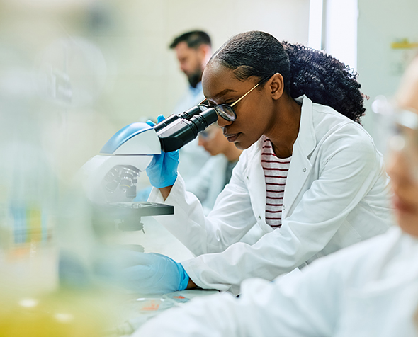 A Black woman in a lab coat peers through the lens of a microscope in a lab.